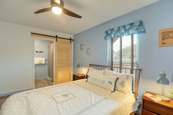 A bedroom with a bed, a nightstand, a ceiling fan, a window with blinds, and a wall-mounted lamp. - Charming Seaside Retreat by Cayucos Pier (Cayucos)