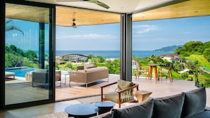 Una sala de estar moderna con un amplio ventanal que da a una terraza con piscina, mobiliario exterior y vistas al océano y colinas.
