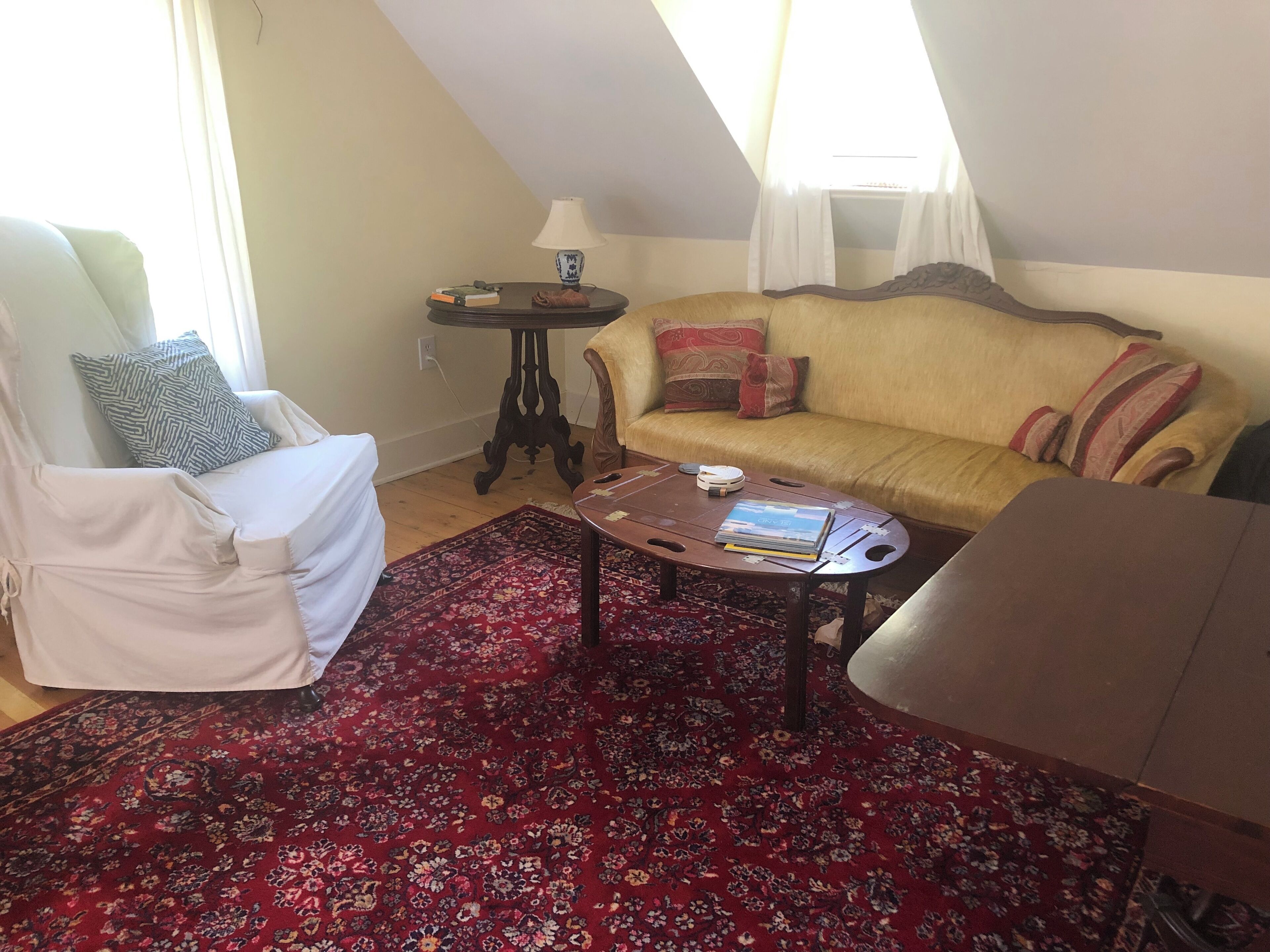 Een gezellige woonkamer met een bank, fauteuil en salontafel, met een rood, patroonrijk vloerkleed en een klein bijzettafeltje met een lamp.