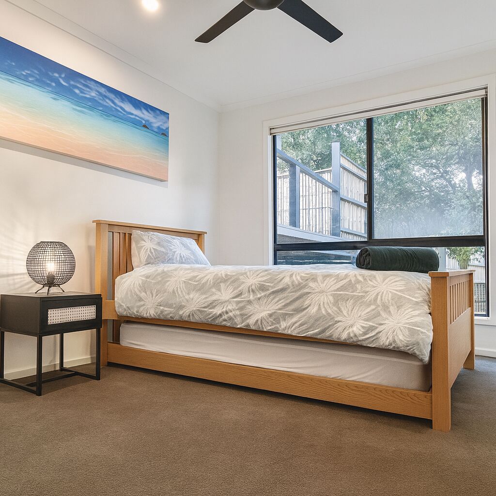 Ein Schlafzimmer mit Bett, Nachttisch, Lampe, Fenster mit Blick auf eine Terrasse und einem Bild an der Wand.