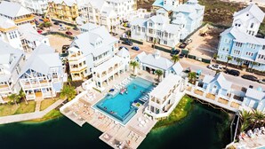 A coastal resort with a central swimming pool surrounded by multiple-story buildings, a pier, and a beach in the background.