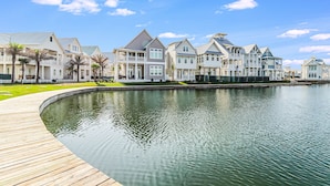 A waterfront property with a wooden boardwalk leading to a calm water body, surrounded by multi-story houses with balconies and a clear blue sky.