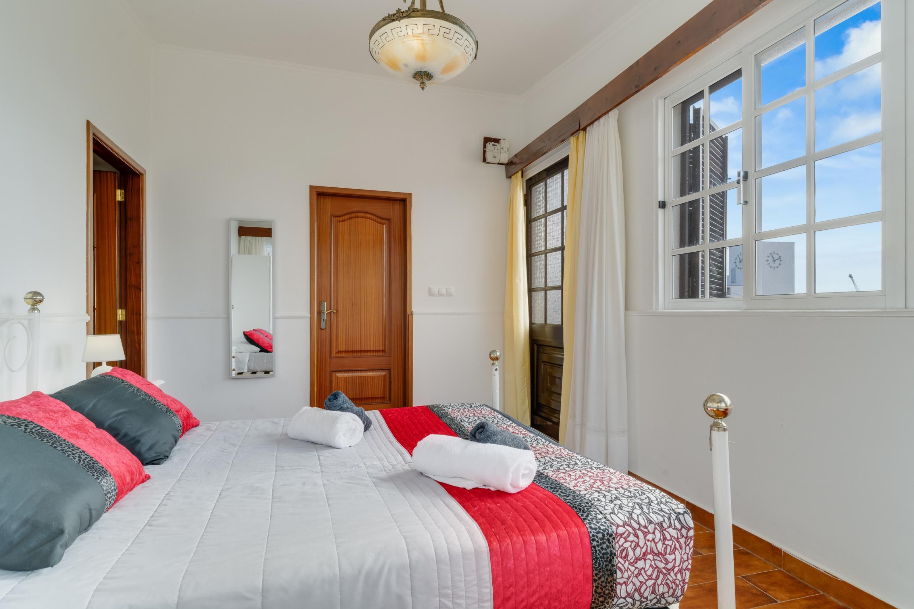 A neatly arranged bedroom with a large bed, a wooden door, a window with curtains, and a ceiling light fixture.