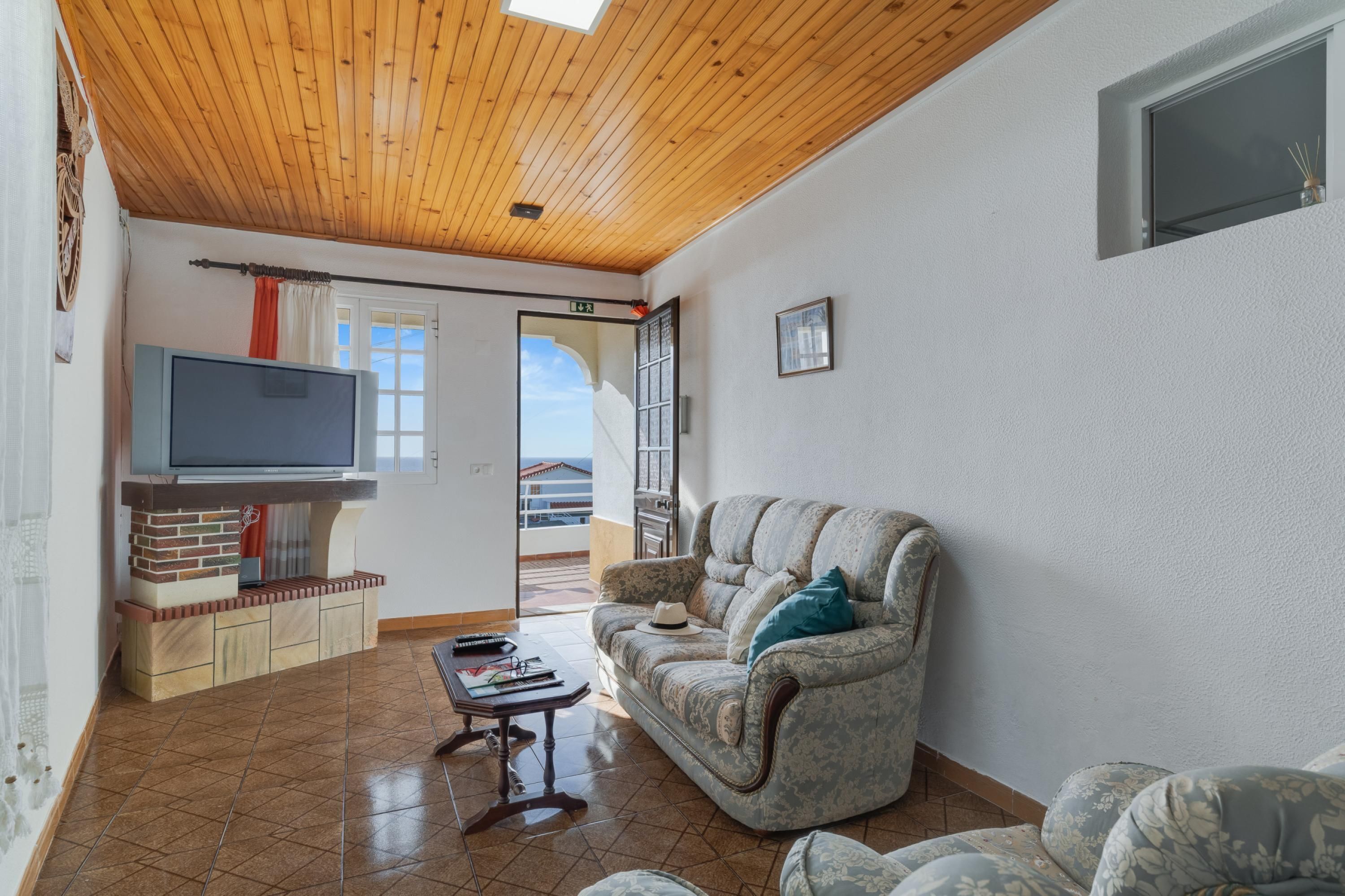A living room with a wooden ceiling, a television on a brick-built entertainment unit, a patterned sofa, and a view through a glass door to the outside.