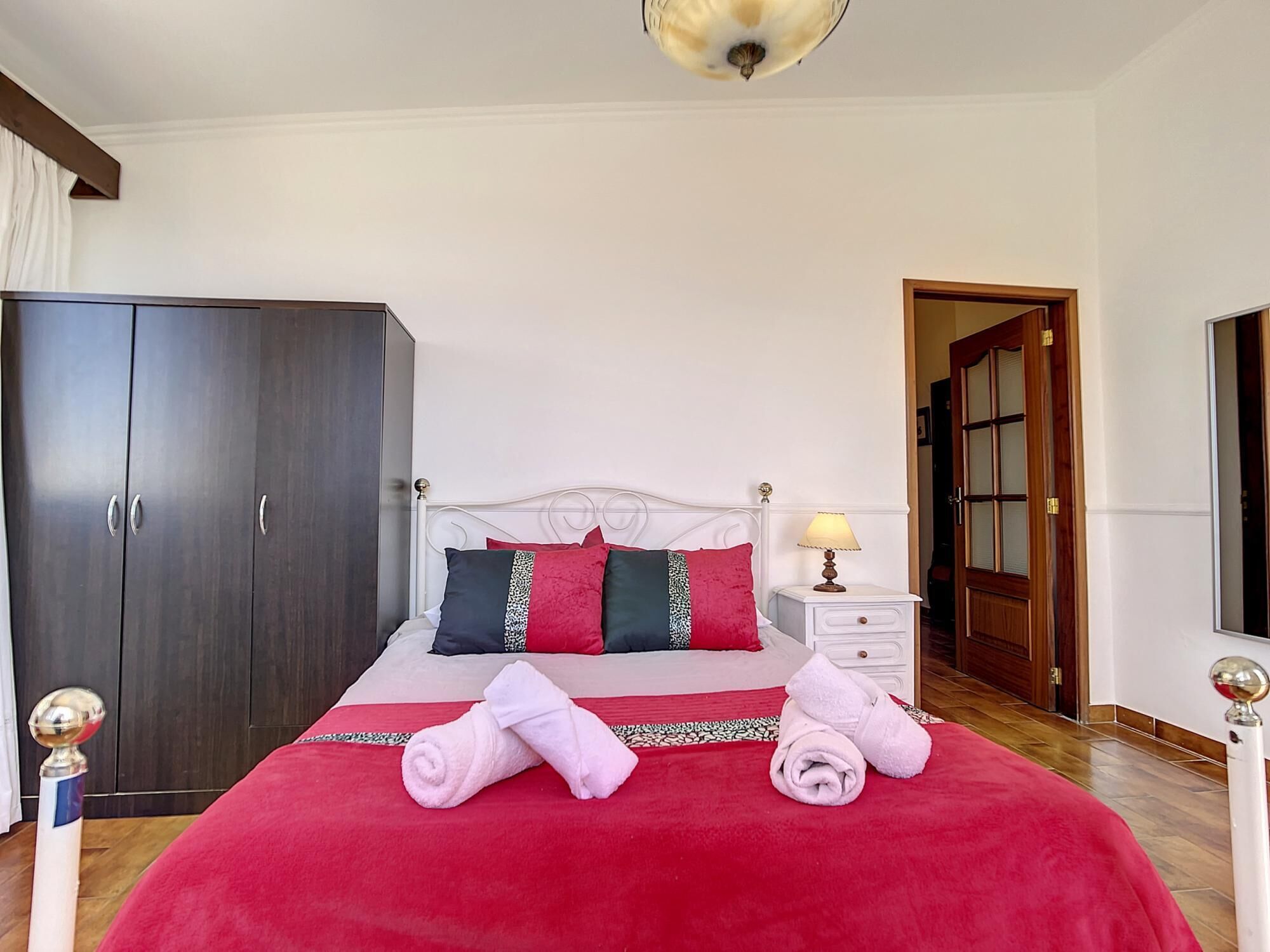 A bedroom with a large bed, a wardrobe, a nightstand with a lamp, and a door leading to another room.