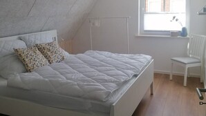 A bedroom with a white bed, patterned pillows, a white radiator, a window with a view of a brick building, and a sloped ceiling. - 2 Bedroom Apartment in Bredstedt (Bredstedt)