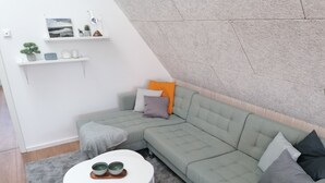 A modern living room with a green sectional sofa, a white coffee table, and a grey carpet. There are shelves with decorative items and a clock on the wall. - 2 Bedroom Apartment in Bredstedt (Bredstedt)