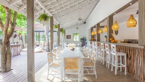 Un área de comedor al aire libre cubierta, con mobiliario blanco, plantas colgantes y techo de madera.