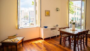 A room with a wooden floor, a dining table with chairs, a white cabinet, a window with a view of buildings, and a basket with flowers. - [Luxury Savona] Cruises 1 min • Free WiFi • A/C (Savona)
