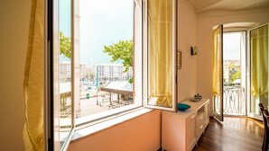 A room with a large window, a white bedside table, and a view of a cityscape. - [Luxury Savona] Cruises 1 min • Free WiFi • A/C (Savona)