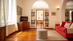 A living room with a red sofa, a round ottoman, wooden flooring, a bookshelf, a TV, and a door leading to another room. - [Luxury Savona] Cruises 1 min • Free WiFi • A/C (Savona)