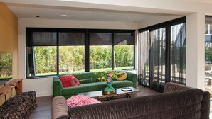 Una sala de estar moderna con un sofá verde, un sofá marrón, una mesa de centro y ventanas amplias con persianas negras.