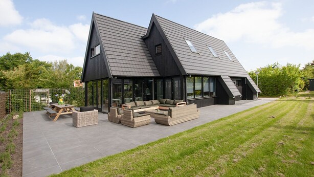 Una casa moderna con techo inclinado, ventanas amplias y un patio con mobiliario exterior.