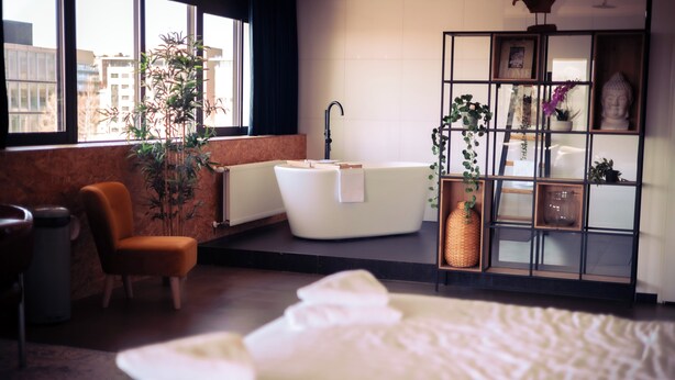 Un baño moderno con una bañera independiente, una silla de madera, un ventanal grande y una planta.