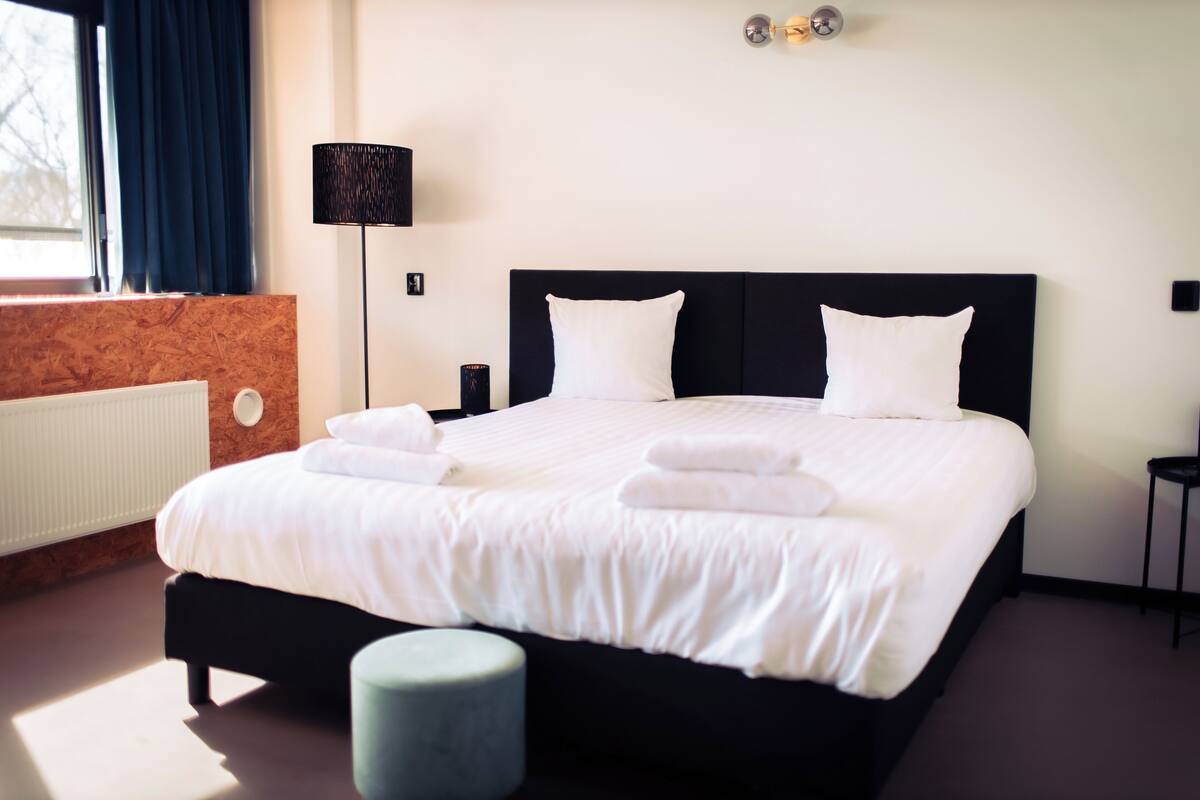 Un lit bien fait, avec du linge de lit blanc et des oreillers, un pouf rond, une lampe sur pied et un radiateur.