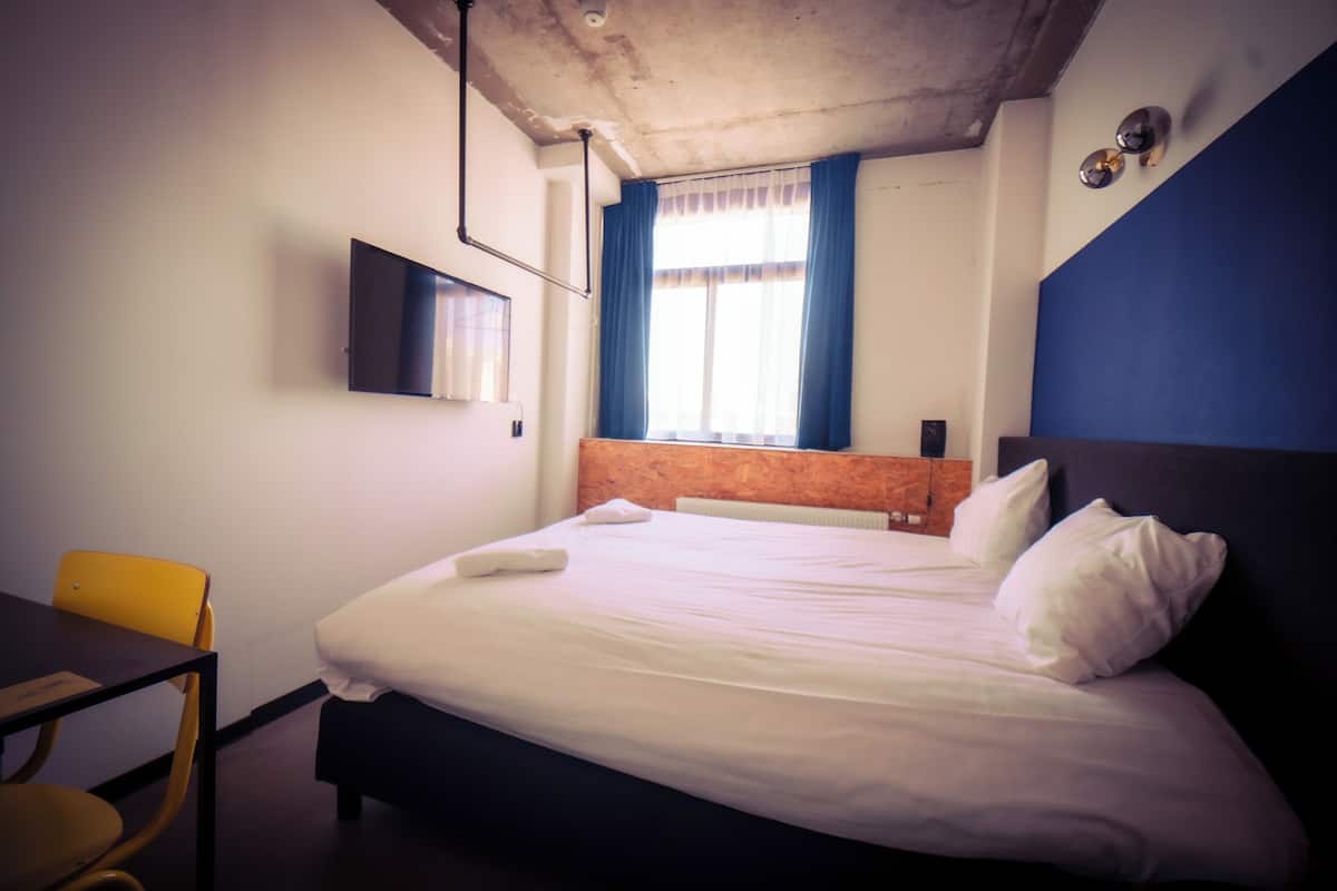 Une chambre d’hôtel comprenant un lit, un bureau, une chaise, un téléviseur fixé au mur et une fenêtre avec des rideaux.