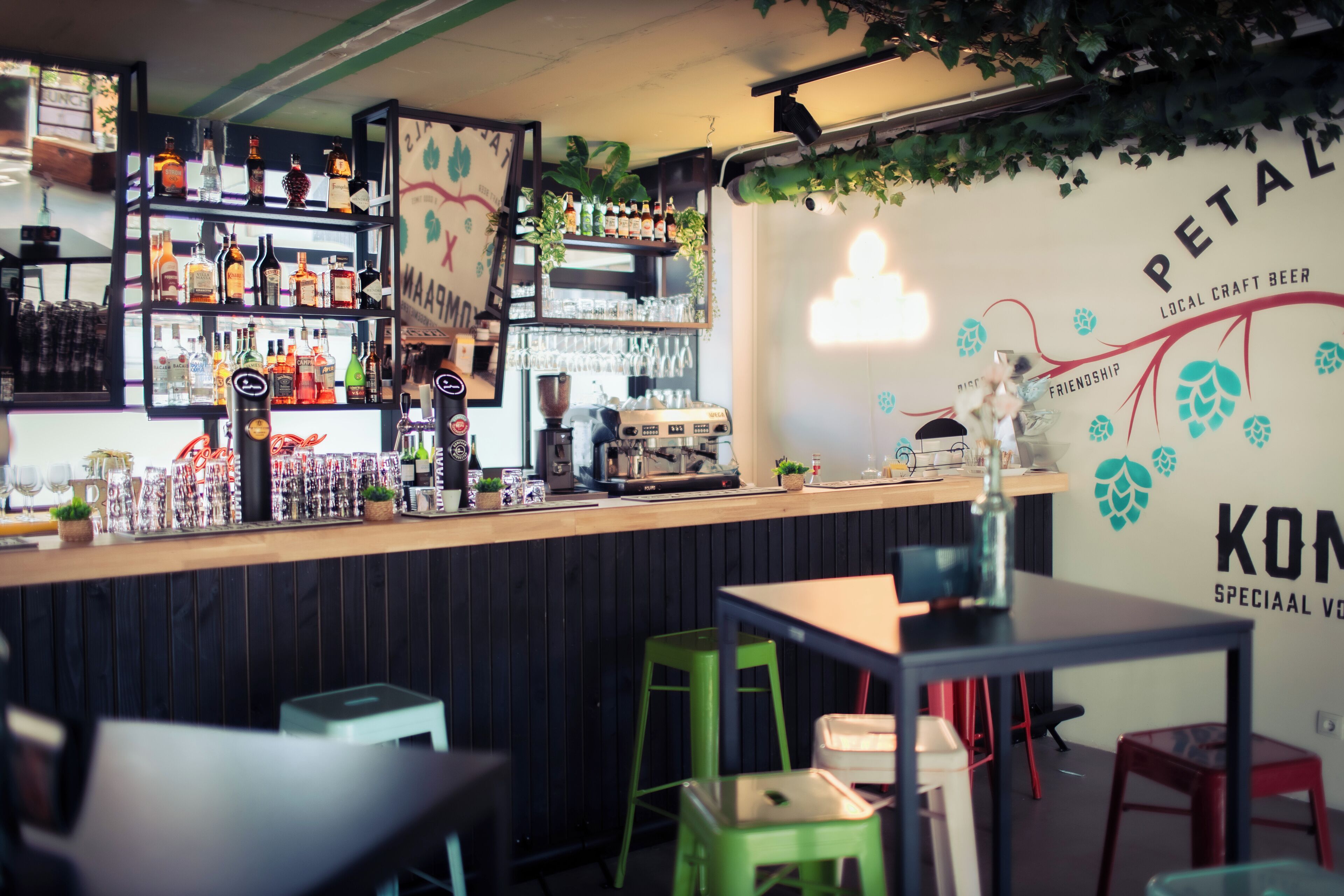 Zona de bar con estantes llenos de botellas y vasos, una barra con una cafetera y una pared con un logotipo y texto.
