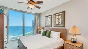A hotel room with a large bed, a ceiling fan, a nightstand with a lamp, and a view of the ocean through large windows.