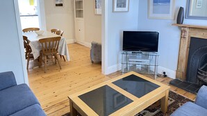 A living room with a TV on a glass stand, a sofa, a dining table with chairs, and a fireplace. - 37 Trenwith Place - a cottage that sleeps 5 guests in 3 bedrooms (St Ives)
