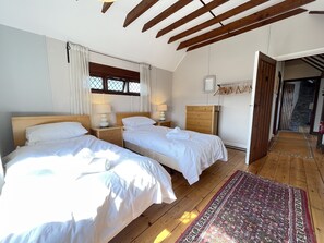 A bedroom with two beds, wooden floors, a patterned rug, a window with curtains, and a wooden ceiling.