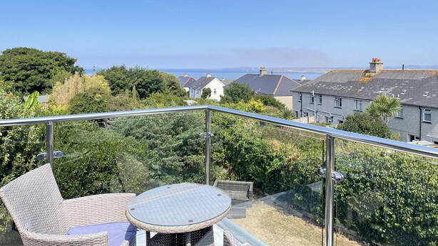 A balcony with a glass railing, wicker furniture, and a view of houses and the sea. - Stratton Vow - a true 1920s Arts and Crafts house! (St Ives)