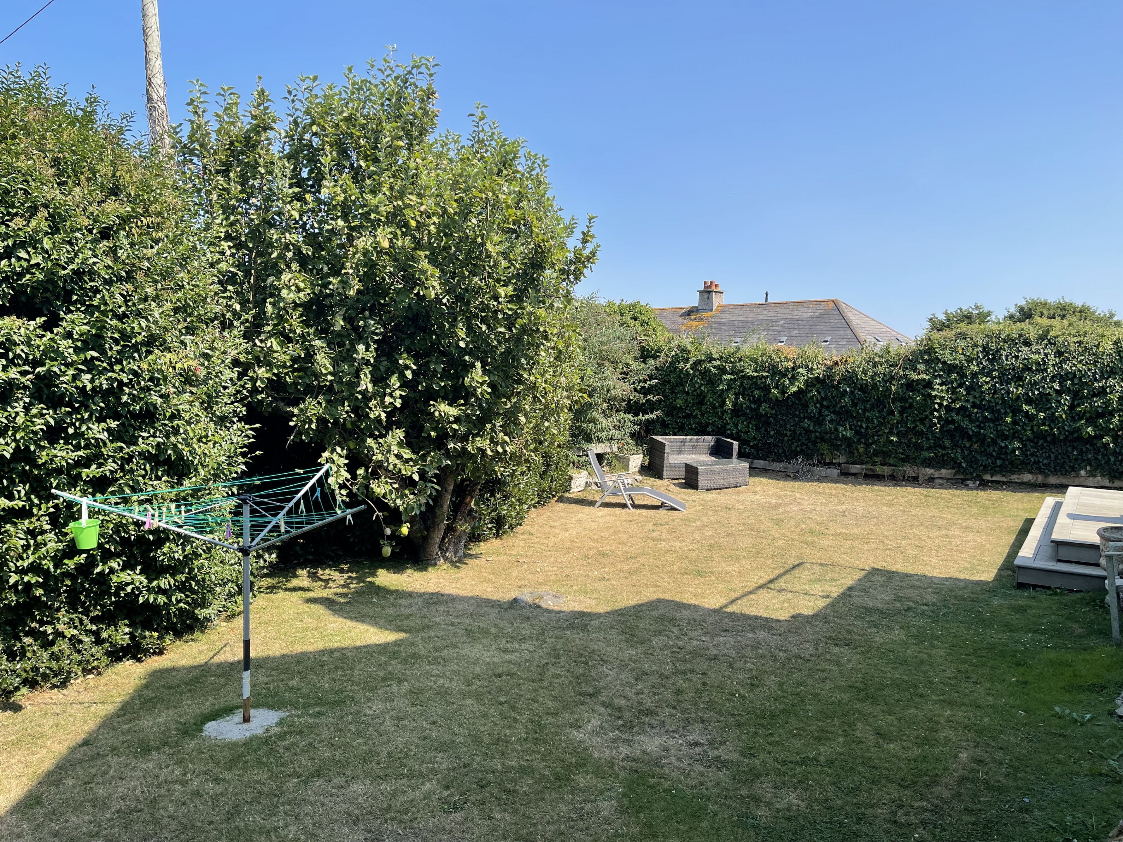 Un jardin avec une pelouse, une terrasse dotée de meubles d’extérieur et une haie.