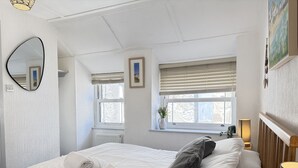 A bedroom with a bed, a bedside table with a lamp, a window with blinds, and a wall-mounted mirror. - Treholme - a cottage that sleeps 6 guests in 3 bedrooms (St Ives)