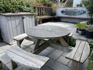Eine Holzterrasse mit einem runden Tisch und Bänken, umgeben von Topfpflanzen und einer Zierwand.