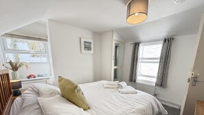 A neatly made bed with a pillow, a bedside table with a lamp, a mirror, and a window with curtains. - Treholme - a cottage that sleeps 6 guests in 3 bedrooms (St Ives)