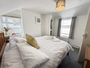 A neatly made bed with a pillow, a bedside table with a lamp, a mirror, and a window with curtains. - Treholme - a cottage that sleeps 6 guests in 3 bedrooms (St Ives)
