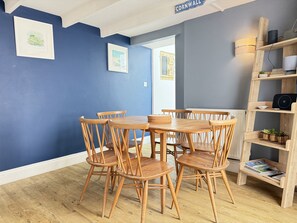 Een eethoek met een ronde houten tafel en stoelen, een boekenkast met boeken en decoratieve objecten, en een blauwe wand met een ingelijst beeld.