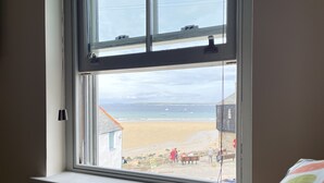 Ein Zimmer mit einem Fenster mit Blick auf einen Strand, einem Heizkörper unterhalb des Fensters und einem Bett mit einem Kissen mit Punkten.