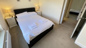A neatly made bed with white linens and towels, two bedside tables with lamps, and a door leading to another room. - Harrys Hideout - a cottage that sleeps 4 guests in 2 bedrooms (St Ives)