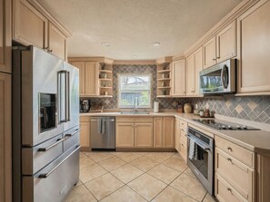A kitchen with light wood cabinets, stainless steel appliances, a tiled backsplash, and a window with a view. - Waterfront Oasis - King Beds • 10min Walk to Beach (Cocoa Beach)