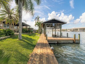 A wooden dock leading to a covered structure by a body of water, with a grassy area and palm trees on the left. - Waterfront Oasis - King Beds • 10min Walk to Beach (Cocoa Beach)