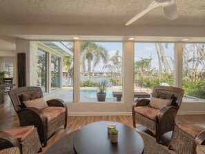 A room with large windows, wicker furniture, a round table, and a view of a pool and palm trees. - Waterfront Oasis - King Beds • 10min Walk to Beach (Cocoa Beach)