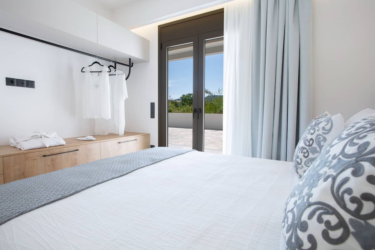 A modern bedroom with a large bed, a wooden dresser, a hanging robe rack, and a sliding glass door leading to a balcony.