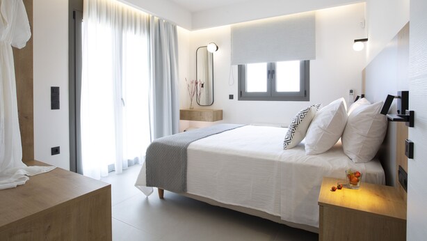 A modern bedroom with a large bed, bedside tables, a mirror, and a window with curtains. - Achillion Suites (Lefkada)
