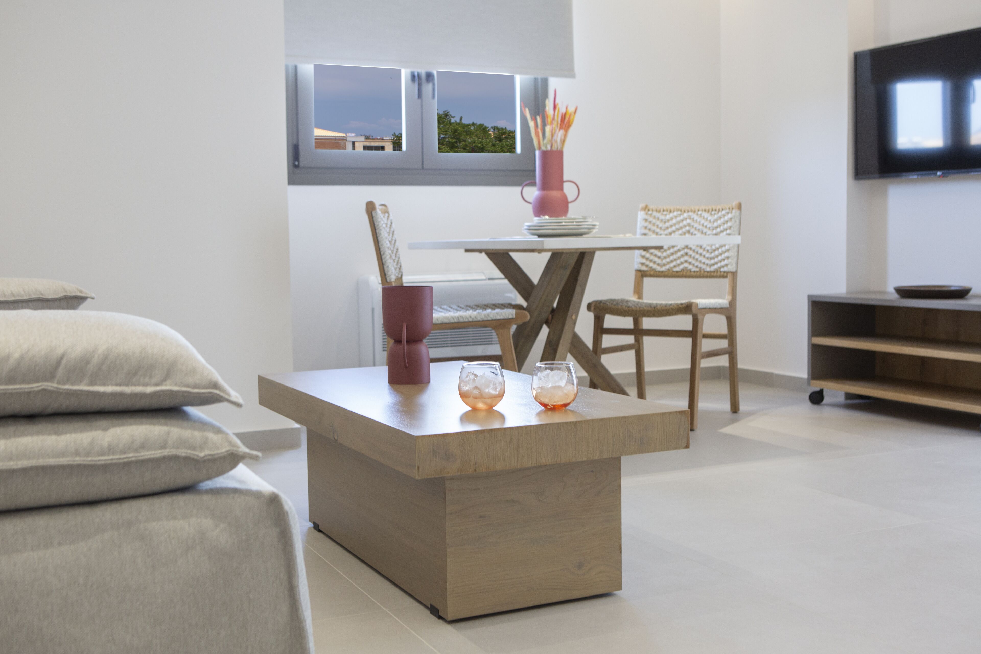 A modern living room with a wooden coffee table, a TV mounted on the wall, and a window with a view of the outside.