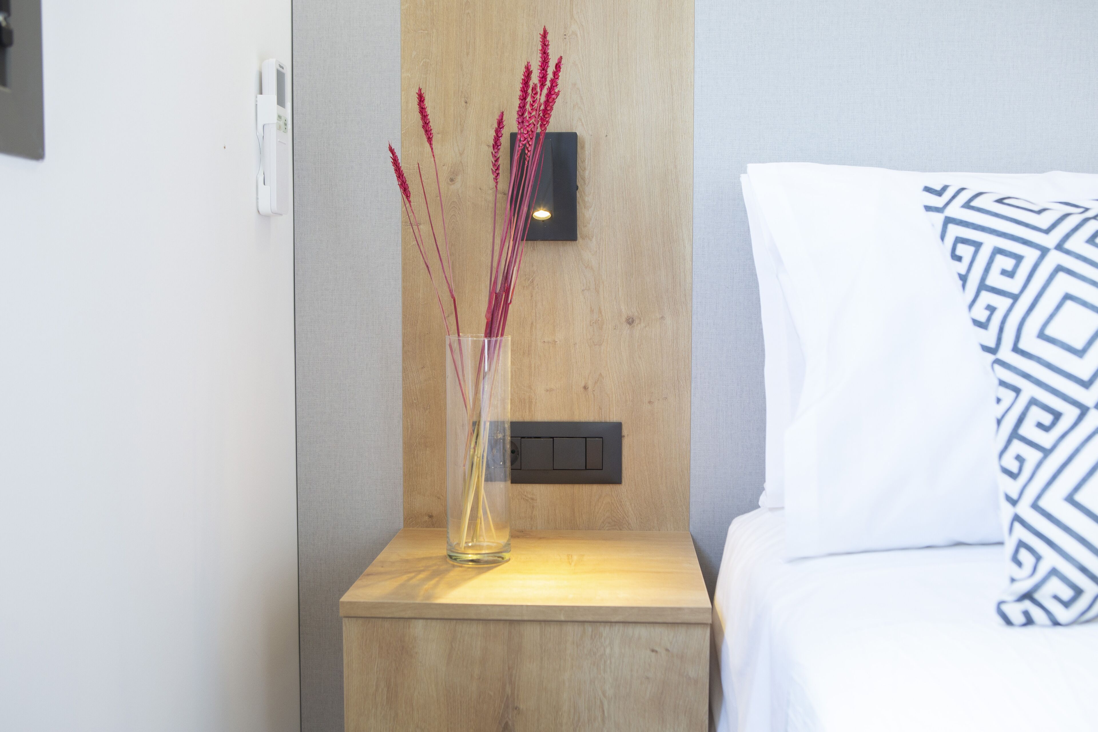 Une table de chevet en bois sur laquelle se trouve un vase en verre transparent contenant des fleurs rouges, une prise de courant et un interrupteur au mur.