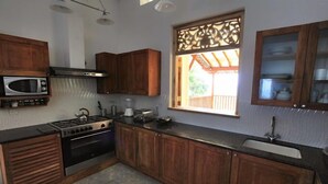 A compact kitchen with wooden cabinets, a stove, a microwave, and a window with a view. - Far Horizons Villa by Angel (Unawatuna)