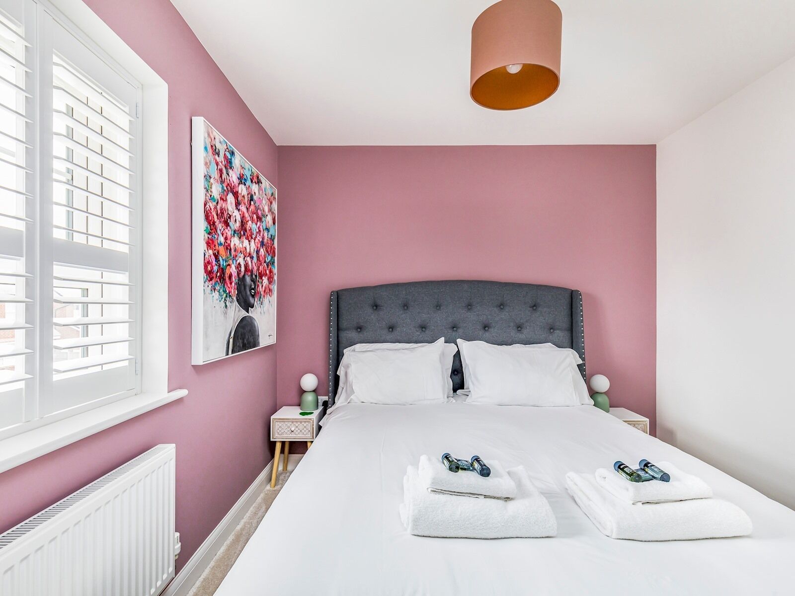 Un dormitorio con pared rosa, cabecero tapizado gris, ropa de cama blanca y un cuadro de flores.