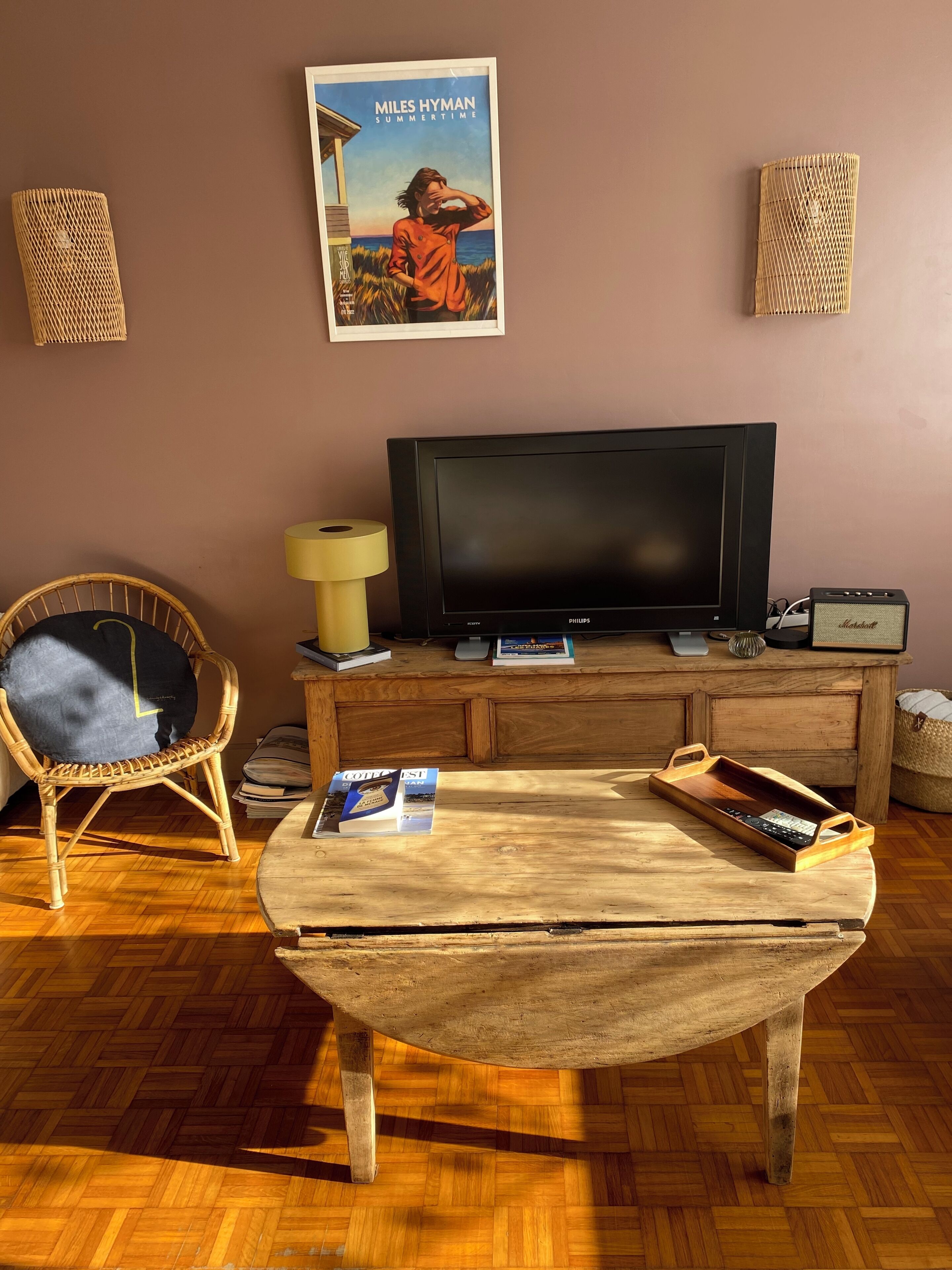 Un salon avec un téléviseur, un fauteuil en osier, une table basse en bois et une affiche accrochée au mur.