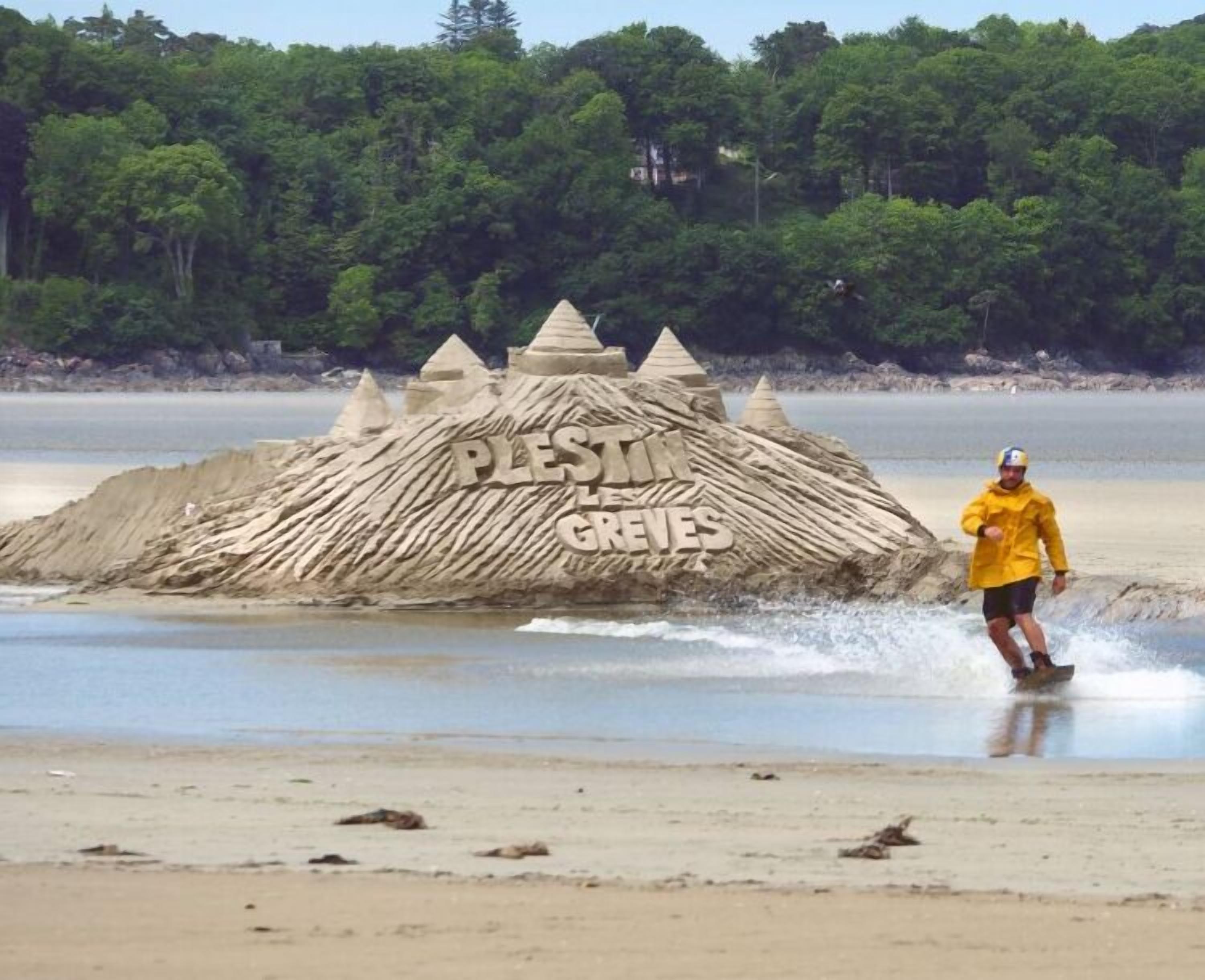 A sandcastle with the words "PLESTI LES GREVES" on it, located on a beach with a person in a yellow jacket riding a wakeboard nearby.