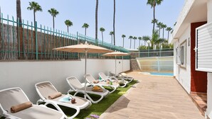 Una fila de tumbonas con toallas, una piscina y palmeras al fondo.