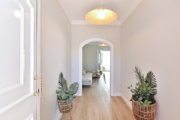 A hallway with a wooden floor, white walls, and a ceiling light fixture. There are two potted plants on either side of the hallway.