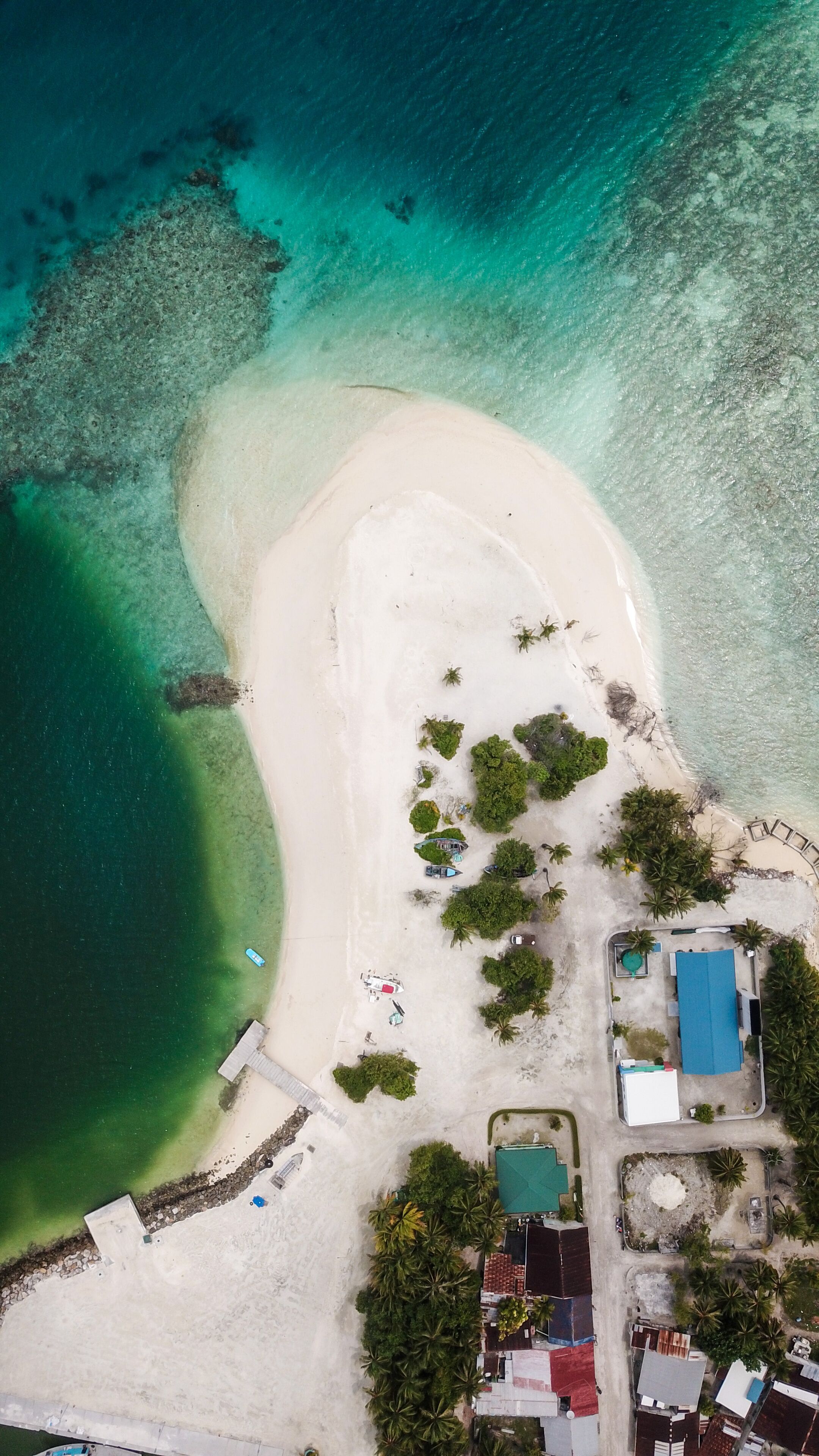 An aerial view of a beach with clear turquoise waters, a sandy shore, and several buildings including a large blue-roofed structure.