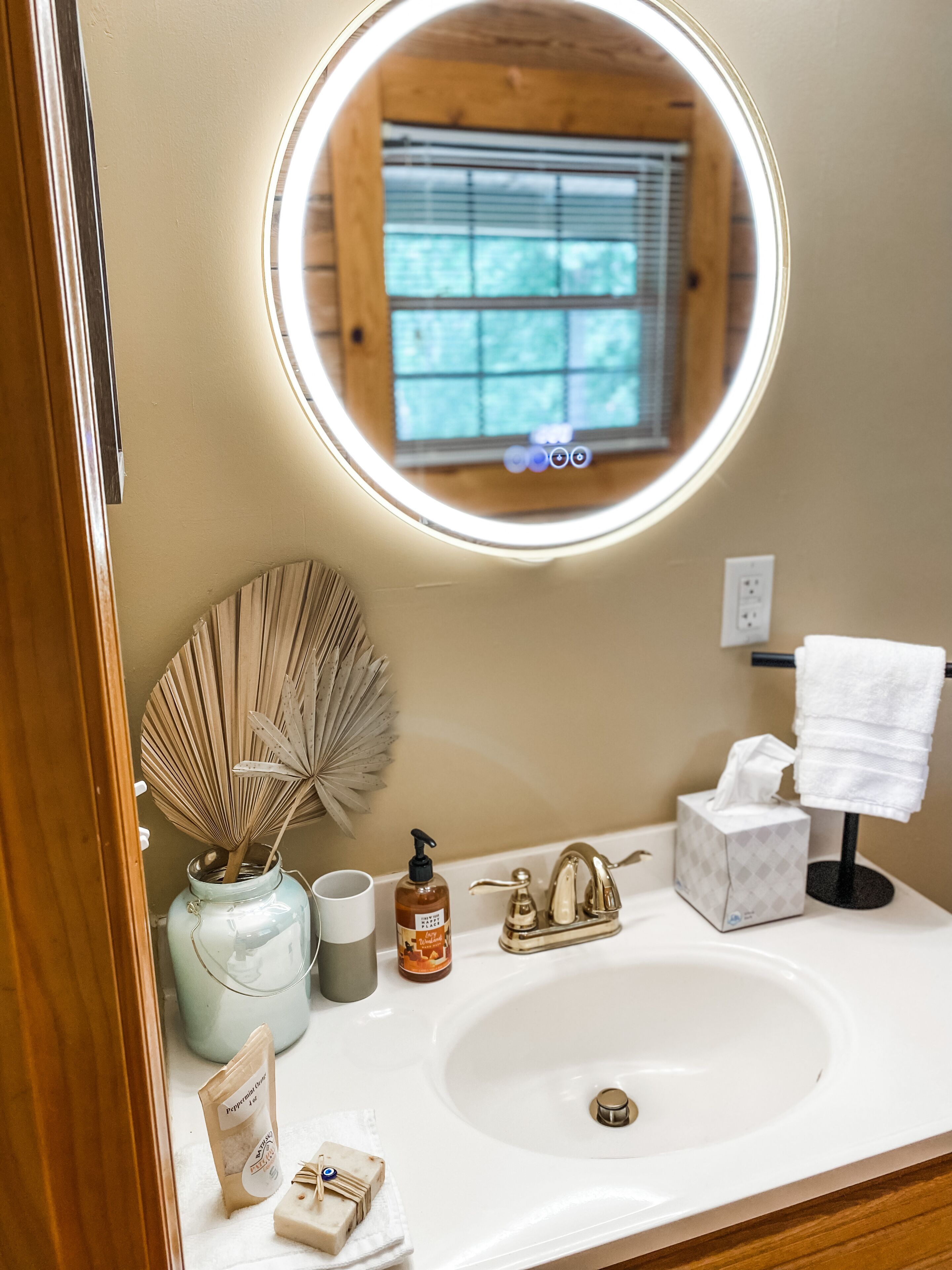 Une salle de bain avec un miroir rond, un lavabo, un porte-serviettes, un ventilateur, un distributeur de savon et une boîte à mouchoirs.