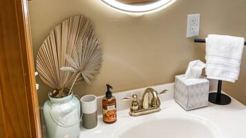 Une salle de bain avec un miroir rond, un lavabo, un porte-serviettes, un ventilateur, un distributeur de savon et une boîte à mouchoirs.