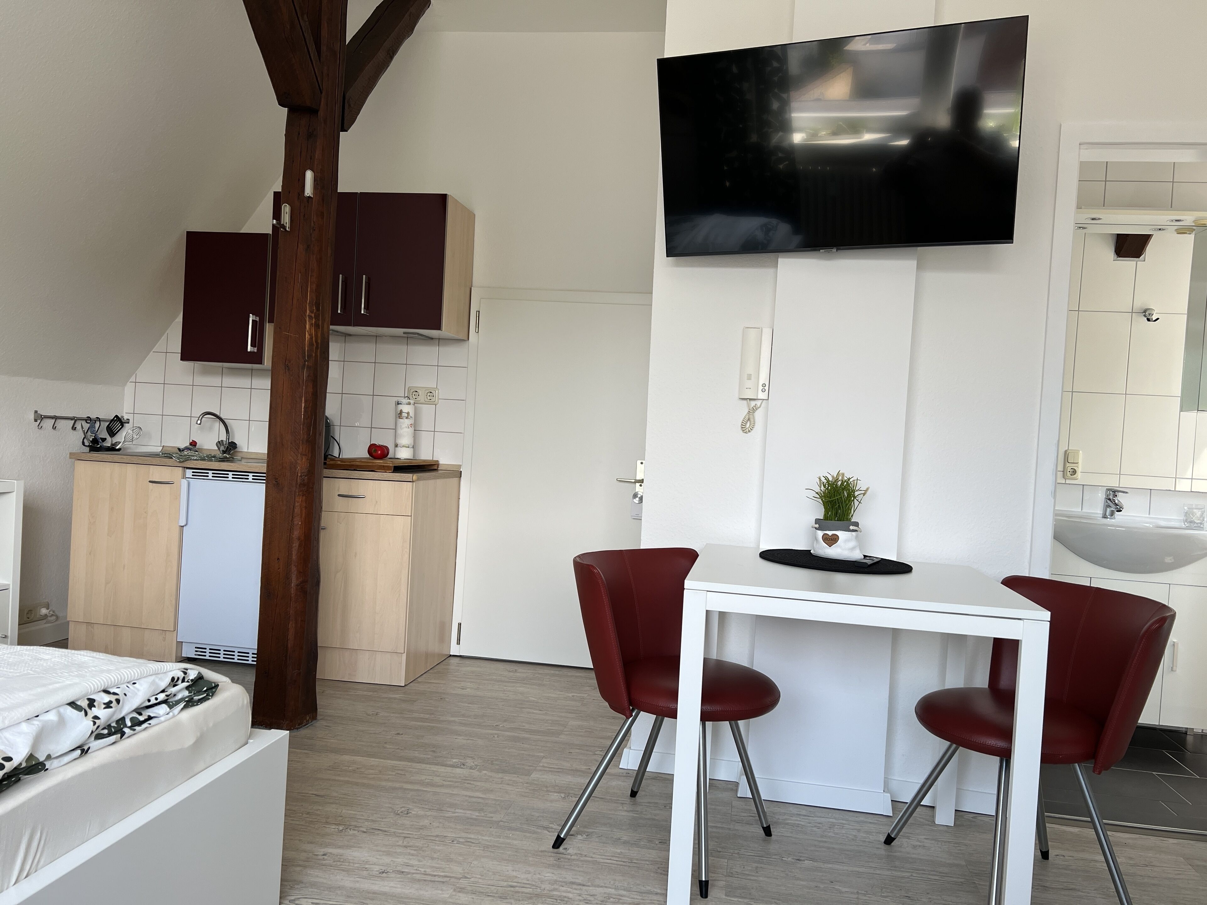 A modern room with a bed, a dining table with two chairs, a TV mounted on the wall, a kitchen area with cabinets and a fridge, and a bathroom with a sink and mirror.